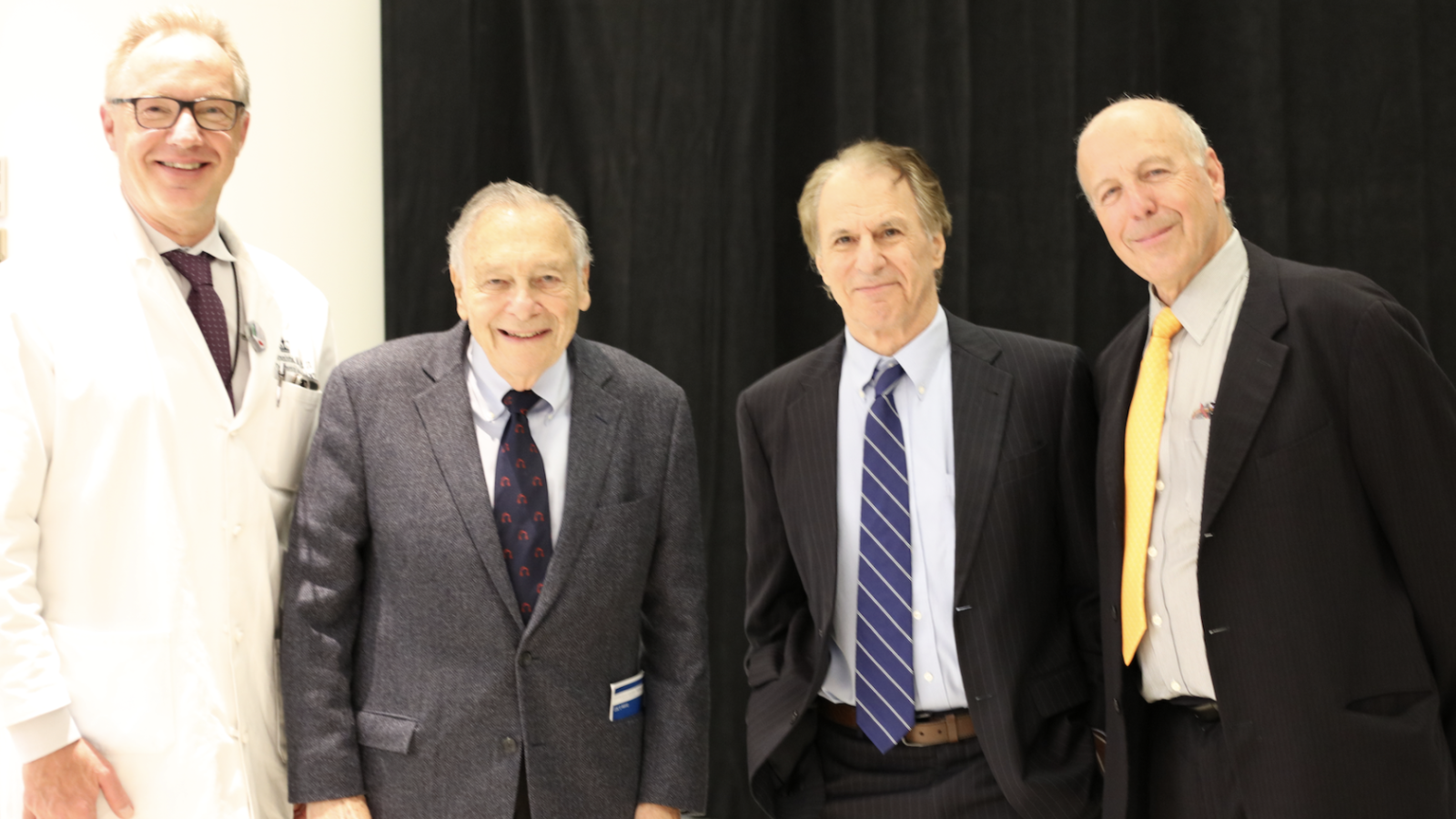 Four men, three in suits, one in doctor's coat pose on stage