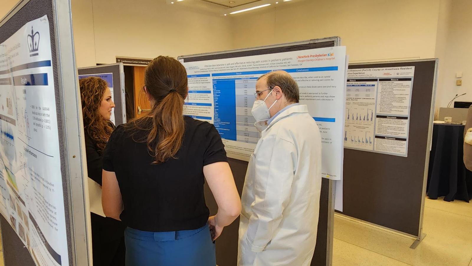 Man in white coat and mask speaking to two women in front of scientific poster