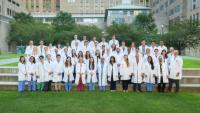 Group of clinicians in white coats, standing on grass and stairs, buildings behind them