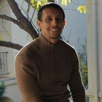 Smiling man in brown sweater in front of a building and a tree