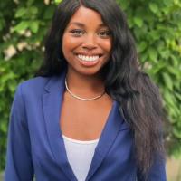 Woman in blue suit and white shirt, smiling, standing in front of greenery